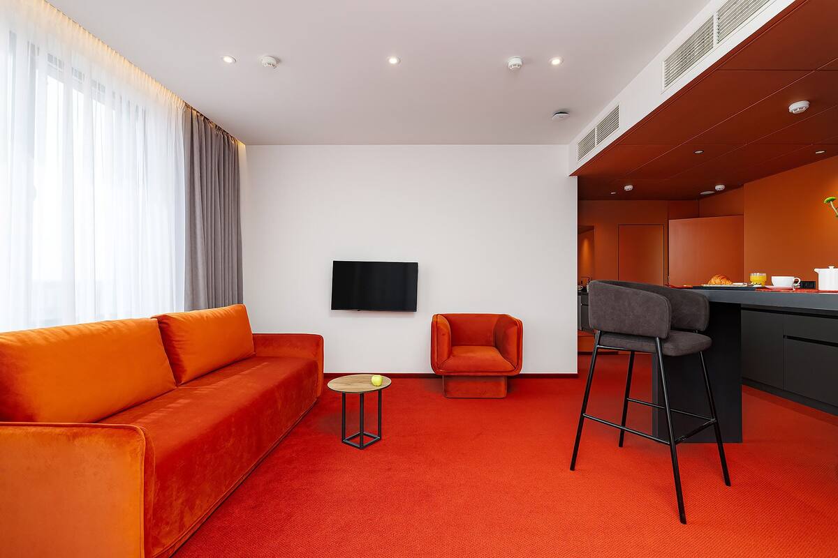 Moderni hotellihuone, jossa on punainen sohva, pieni pyöreä pöytä, televisio ja pieni keittotila.