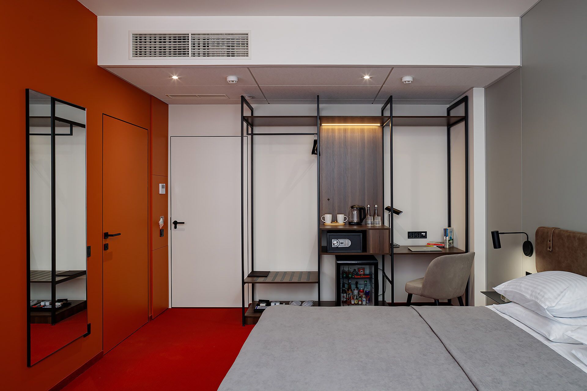 A modern bedroom with a bed, a desk, and a chair. There is a wardrobe with a mirror, a shelf with books, and a small cabinet.