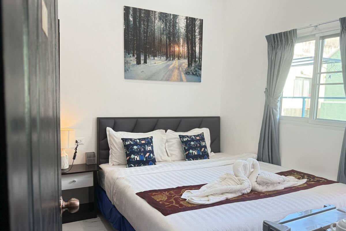 A hotel room with a bed, a nightstand, a lamp, a painting of a snowy forest, and a window with curtains.