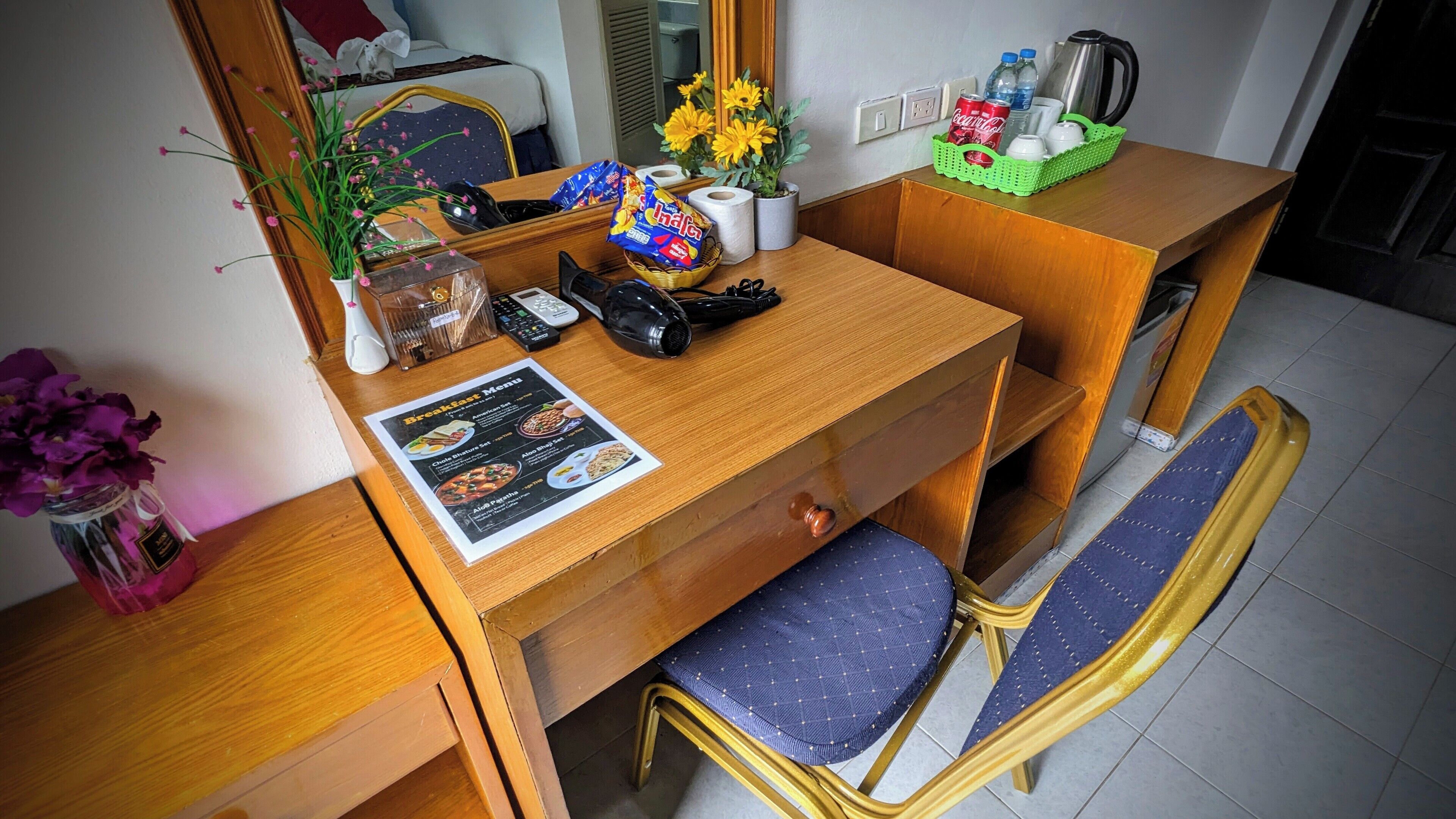 A hotel room with a wooden desk, a blue cushioned chair, a breakfast menu, a vase with purple flowers, a potted plant with yellow flowers, a basket with snacks, and a kettle.