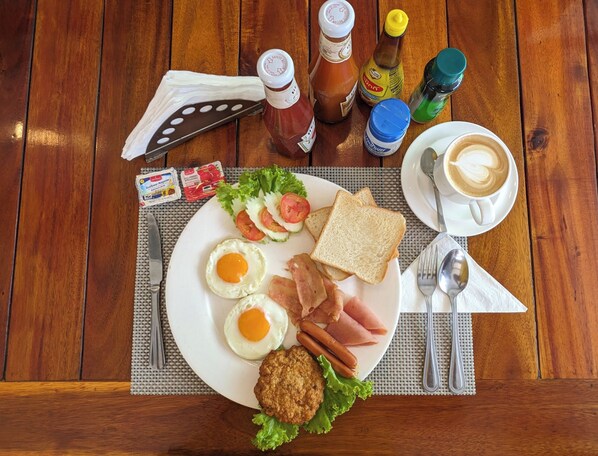 A breakfast plate with eggs, bacon, ham, toast, and a muffin, along with condiments and a cup of coffee on a wooden table. - Heaven House Sapphire Sky (Patong)
