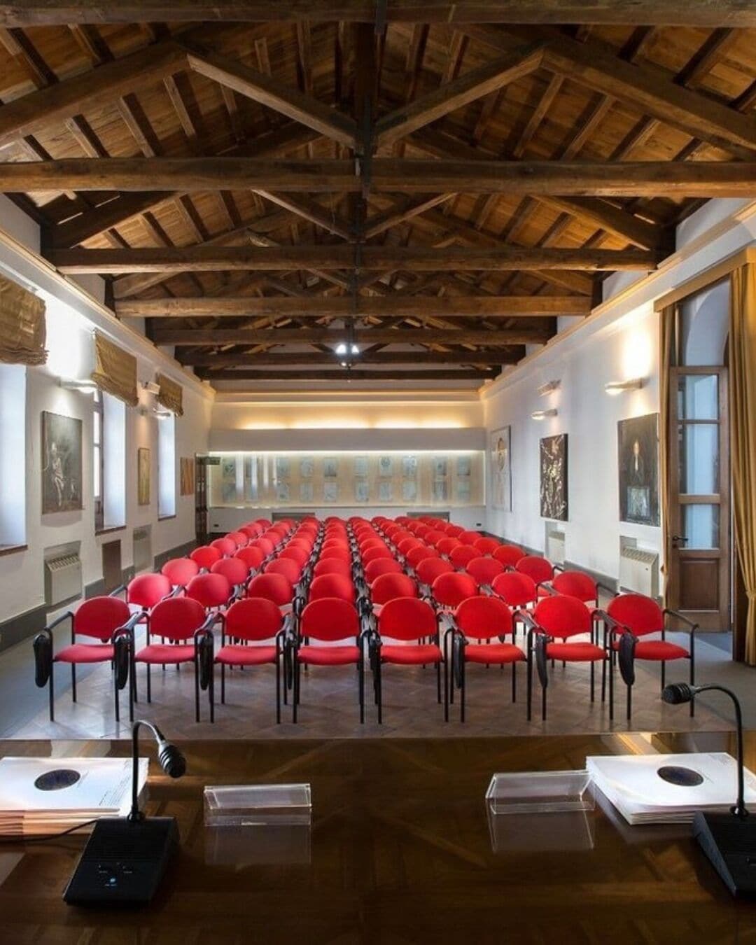 A conference room with red chairs arranged in rows, wooden beams on the ceiling, and artwork on the walls.