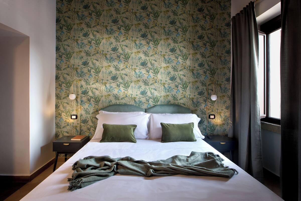 A neatly made bed with a green blanket, a floral wallpaper, and a nightstand with a book and a lamp.
