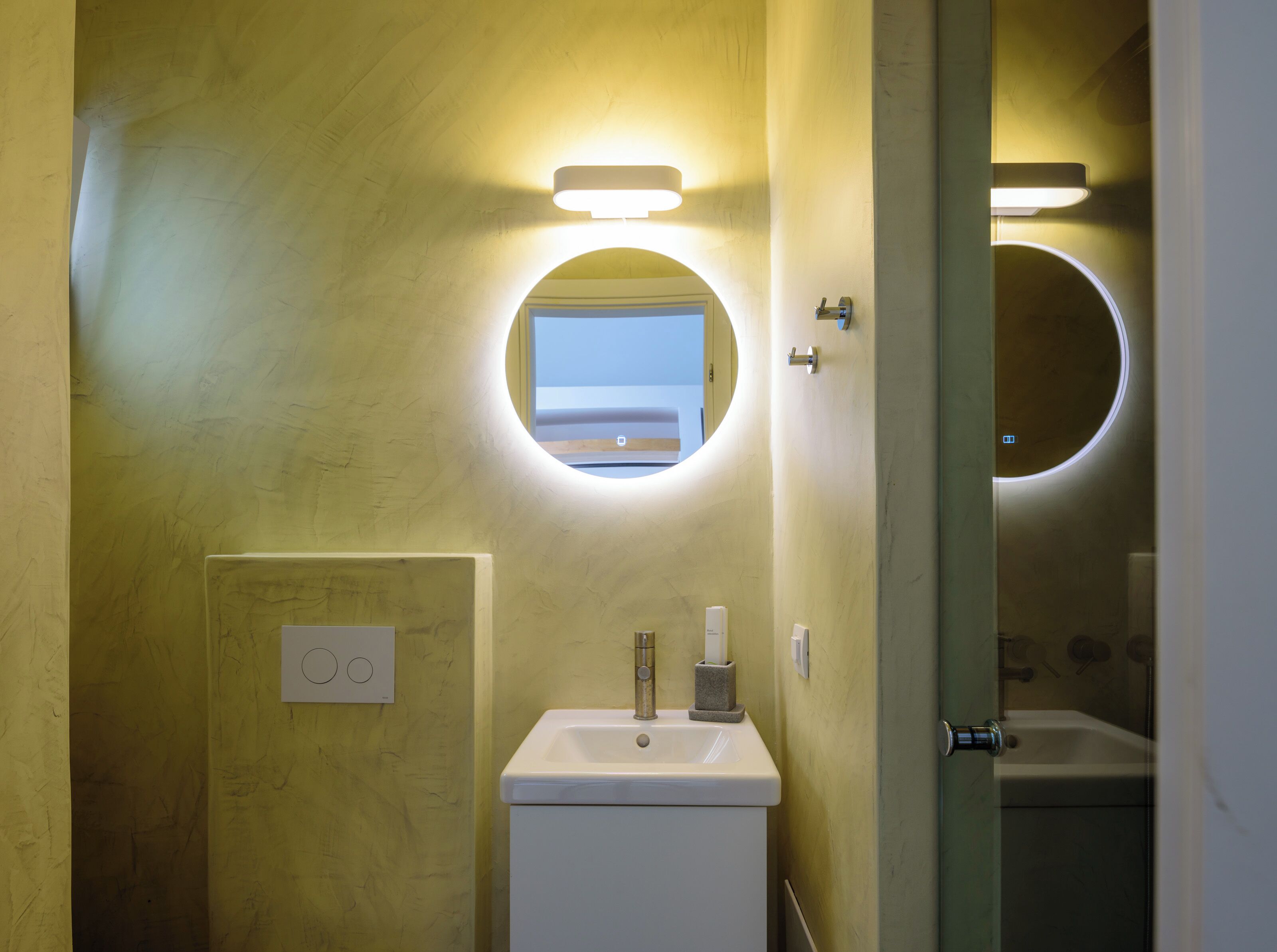 A bathroom with a round mirror, a sink, and a toilet. There is a light fixture above the mirror and a light fixture above the toilet.