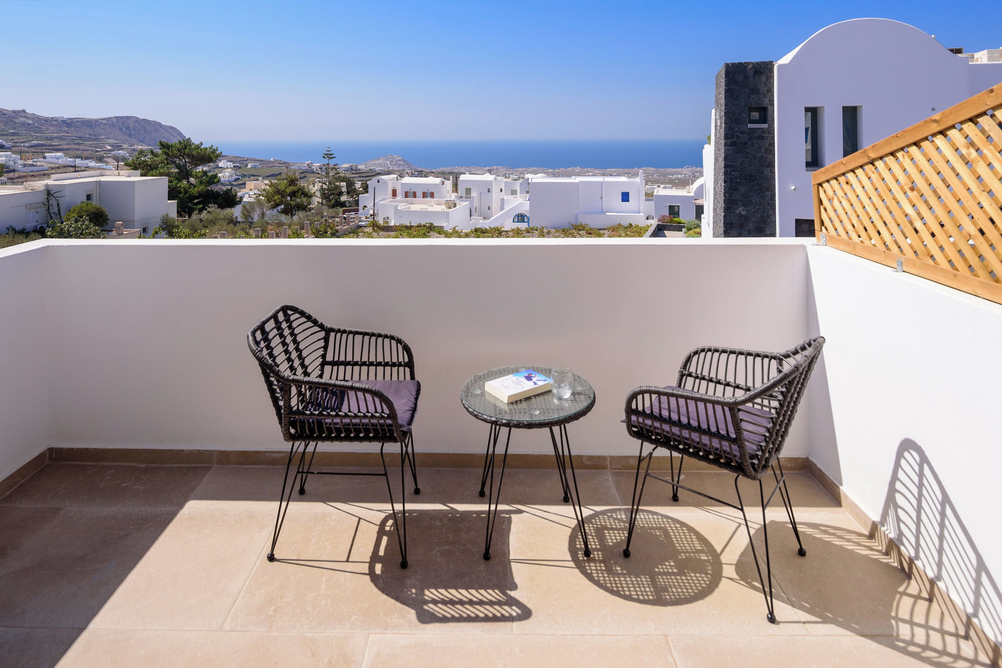 Un balcon avec deux fauteuils en osier et une petite table, donnant sur une ville côtière aux bâtiments blancs et offrant une vue sur la mer.