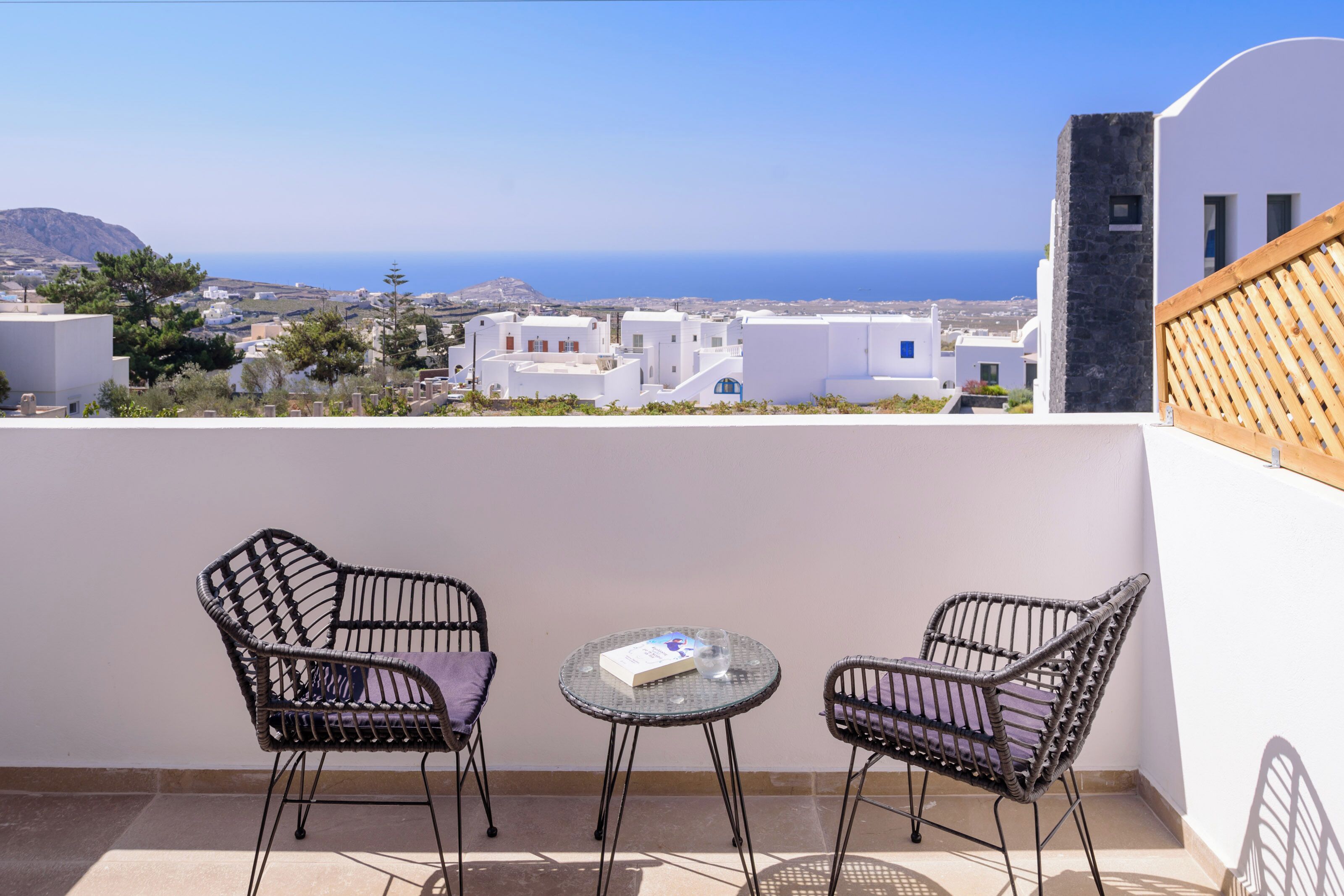 Un balcón con dos sillas de mimbre y una mesita, con vistas a un pueblo costero de edificios blancos y al mar.