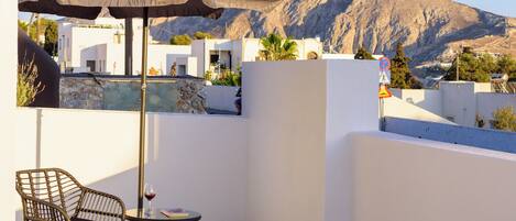 A balcony with a table, chair, and umbrella, overlooking a mountainous landscape with white buildings.