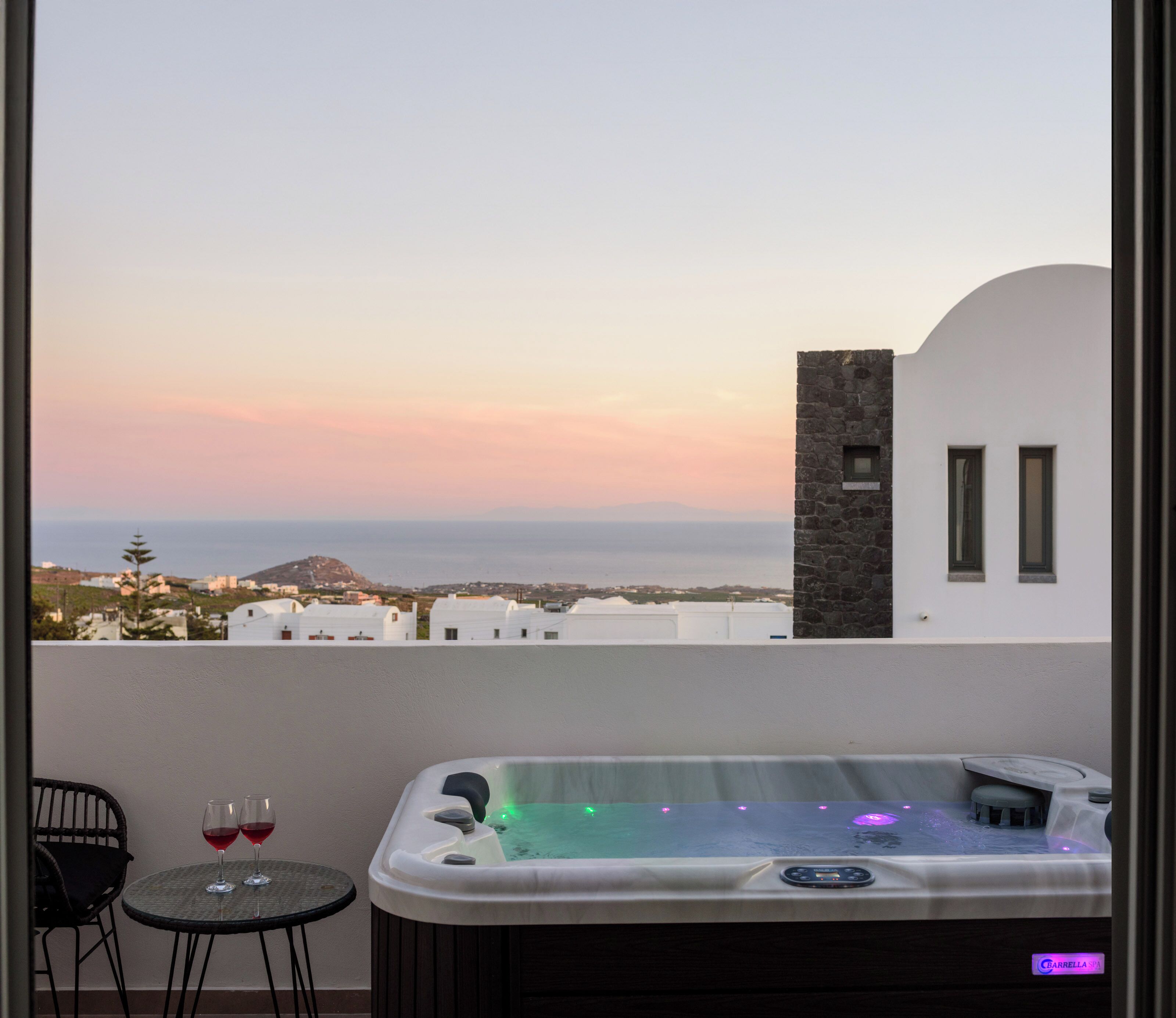 A balcony with a hot tub, two wine glasses, and a view of the sea and buildings.