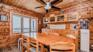 A wooden cabin interior with a dining table, chairs, a ceiling fan, a refrigerator, and a window overlooking a balcony.