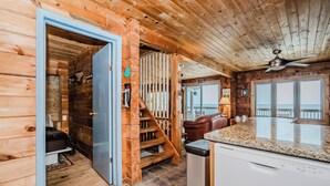A wooden cabin interior with a kitchen area, a dining table, and a view through the window.