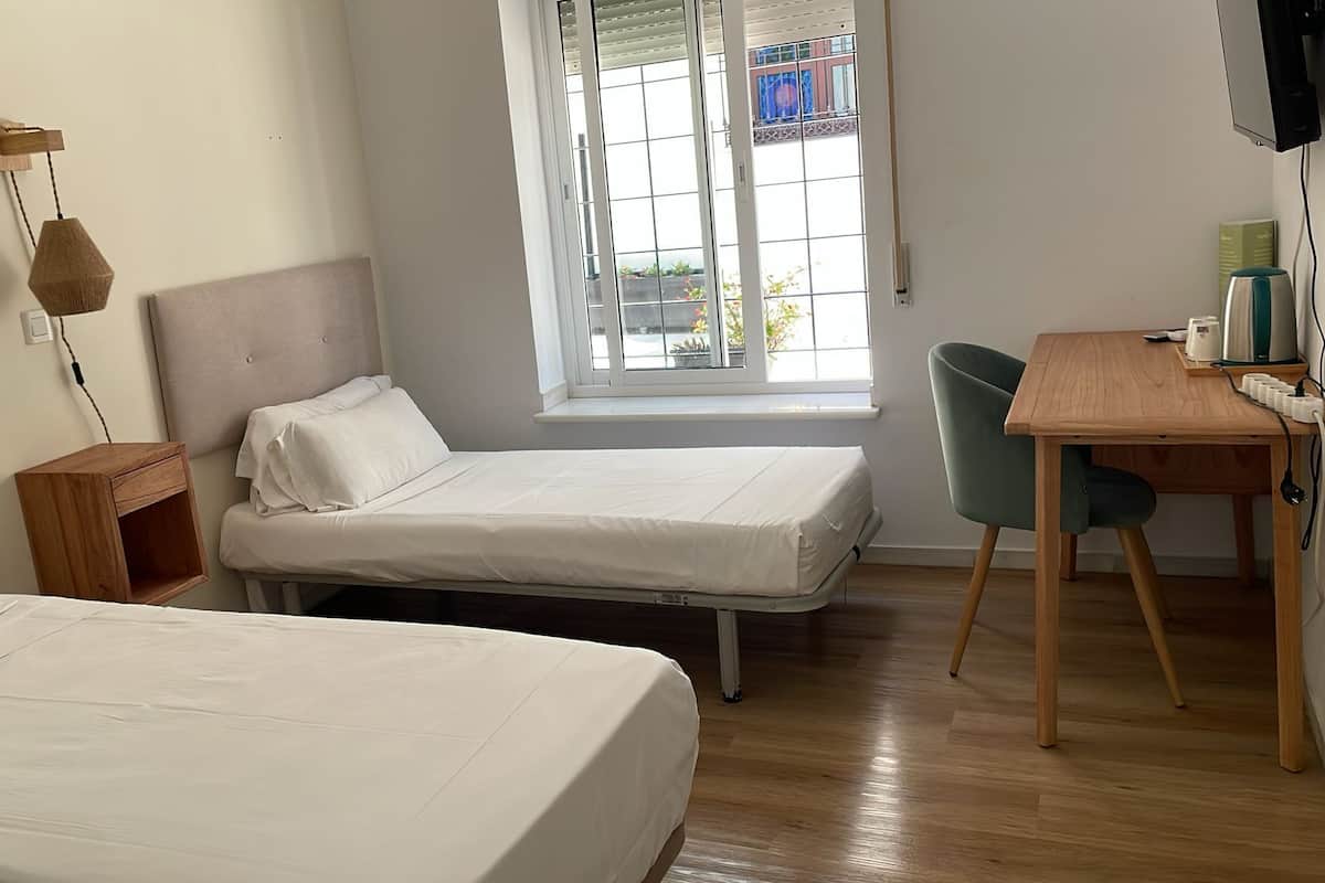 A small, clean room with two single beds, a wooden desk, and a chair. There is a window with a view of the outside.