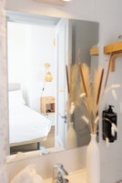 Une chambre d’hôtel avec un lit, une table de chevet avec une lampe, un miroir et un vase contenant des plantes séchées.