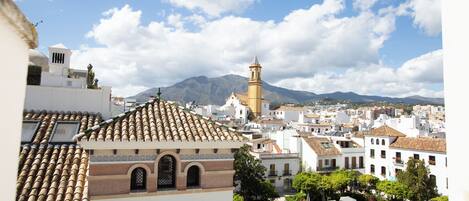 Una plaza de pueblo con una fuente, rodeada de edificios de arquitectura tradicional y una montaña al fondo.