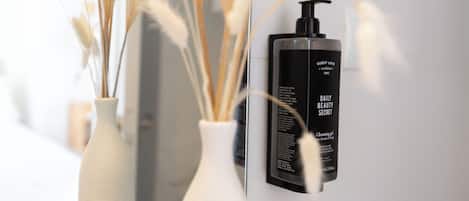 Un baño con un jarrón blanco con ramas secas, una botella negra de loción para huéspedes y un espejo que refleja la habitación.