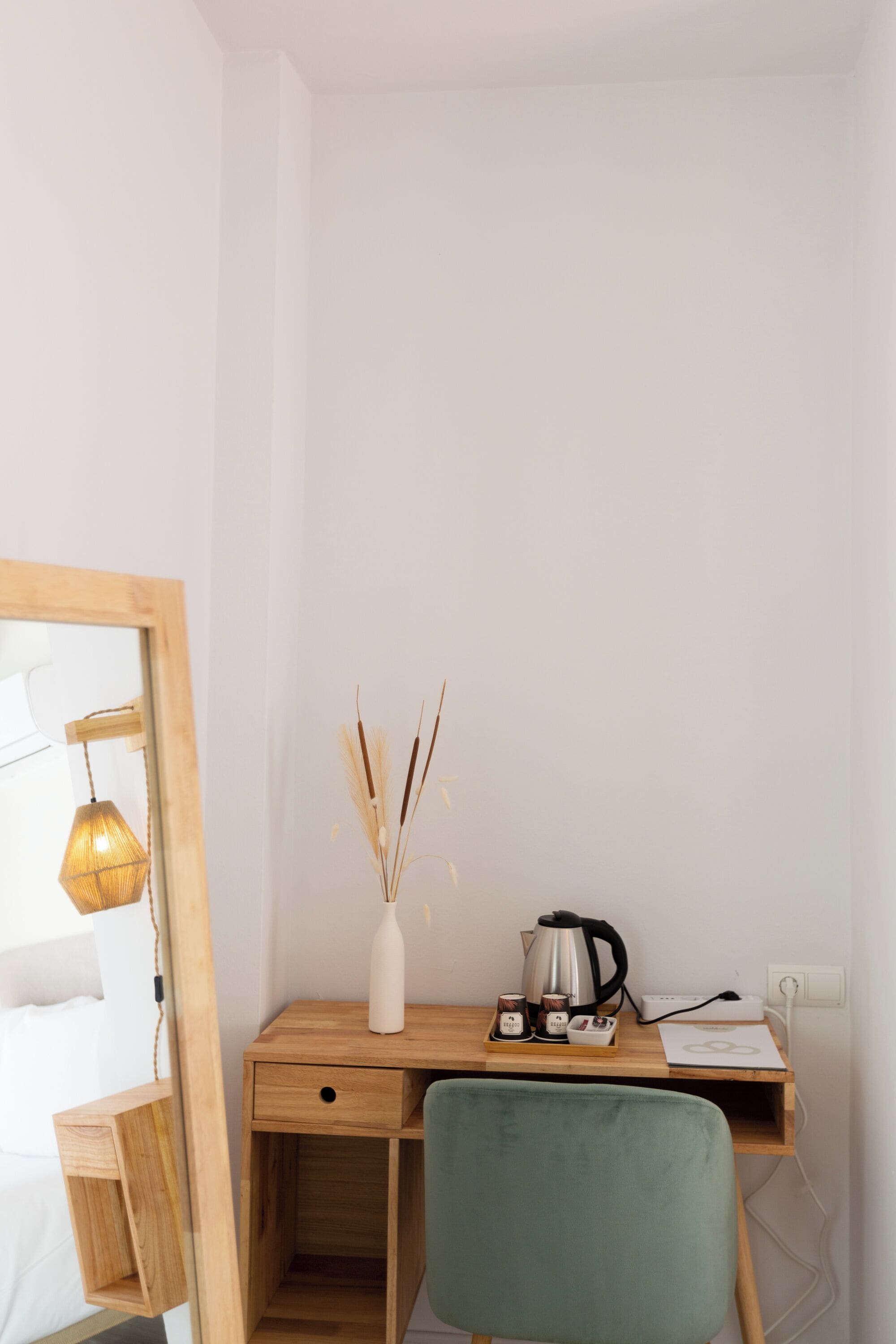 Un petit espace de travail bien rangé, avec un bureau en bois, une chaise verte, un vase blanc contenant des plantes séchées, une bouilloire et quelques objets personnels.
