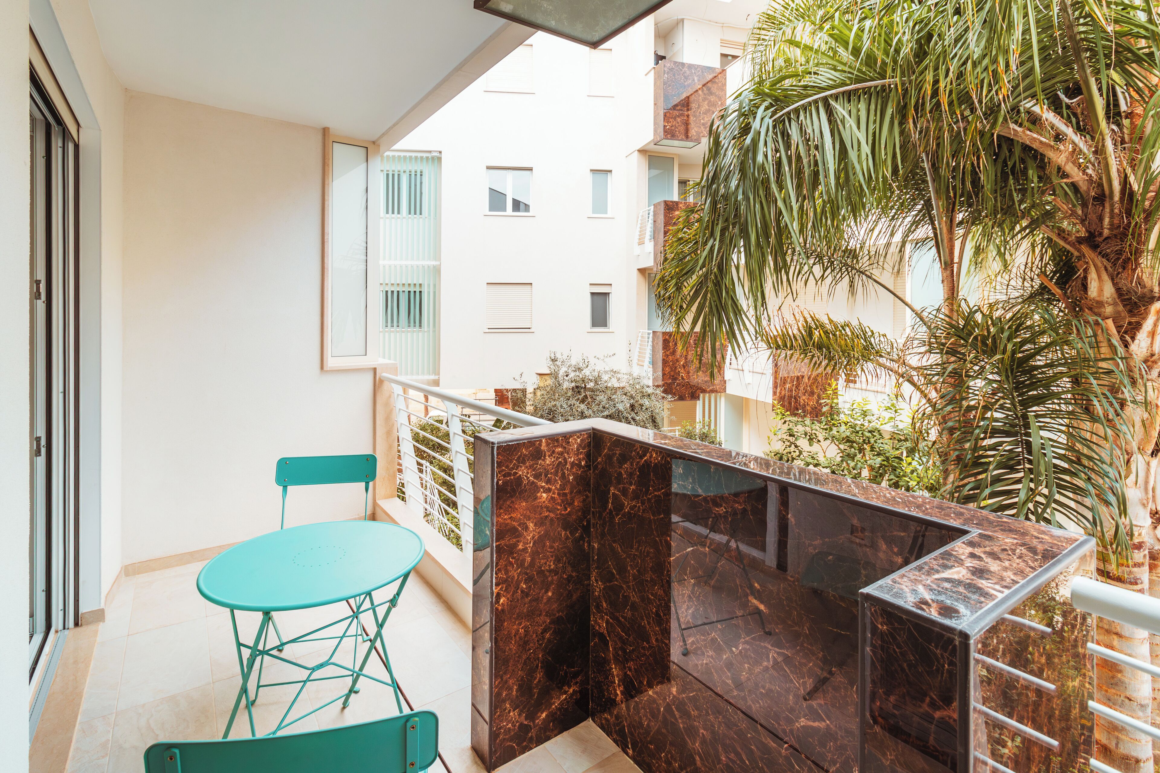 A balcony with a table and chairs, a view of neighboring buildings, and a palm tree.