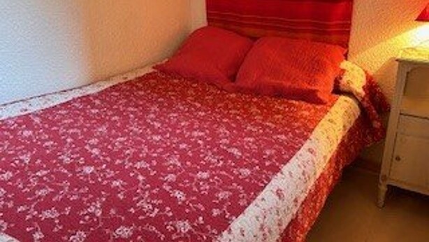 Ein kleines Zimmer mit einem Einzelbett, einer roten Bettdecke mit Blumenmuster, einem Holz-Kopfteil und einem Nachttisch mit Lampe.