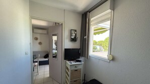 Une petite pièce comprenant un lit, un bureau avec un ordinateur, une télévision et une fenêtre donnant sur des arbres.