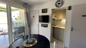 Un coin repas avec une table ronde, un téléviseur fixé au mur et une cuisine visible à travers une porte ouverte.