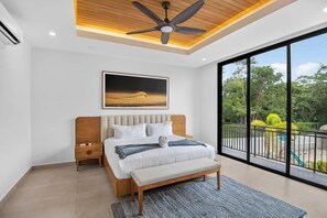Un dormitorio moderno con cama, ventilador de techo, un ventanal y vistas a la vegetación.