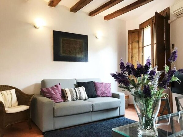 A living room with a grey sofa, a wicker chair, a glass coffee table, and a vase with purple flowers.