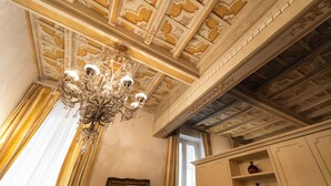 A chandelier with multiple light fixtures, ornate ceiling with floral patterns, and a window with curtains. - Boccaccio 22 - 4 (Roma)