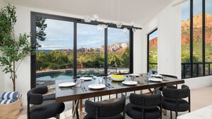 Espaço de refeições com uma mesa grande, cadeiras e vista para uma piscina e montanhas.
