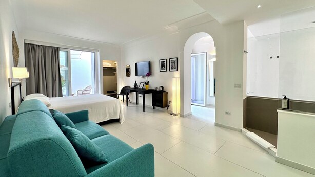 A modern hotel room with a bed, teal sofa, desk, TV, and a view of the sea. - Casa Giuliani Capri (Capri)