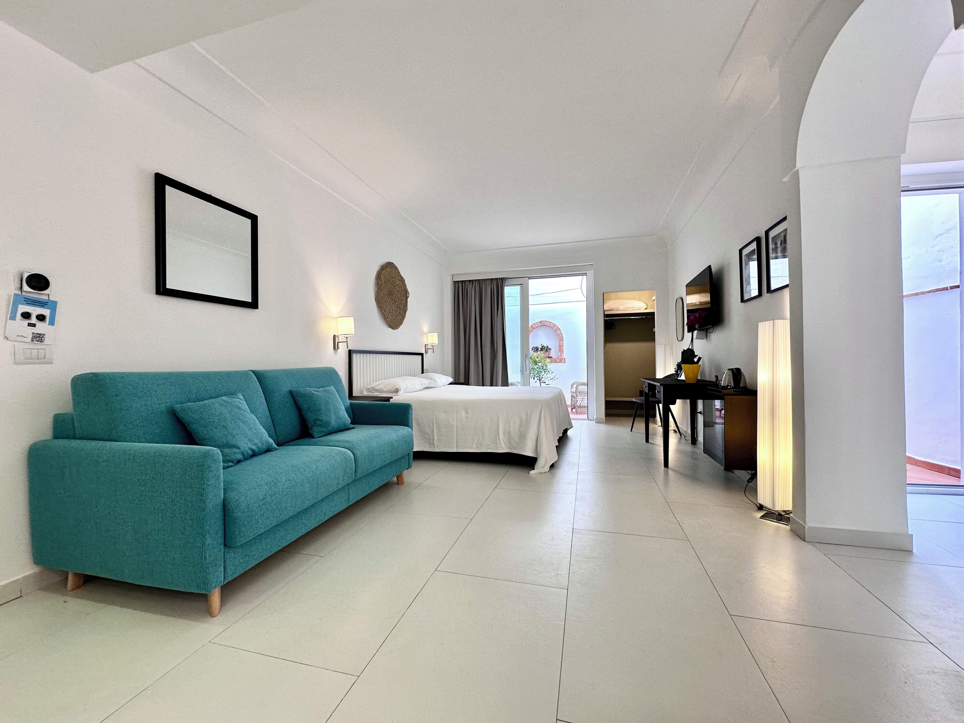 A hotel room with a teal sofa, a bed with white linens, a desk with a lamp, and a view of the sea through a glass door.