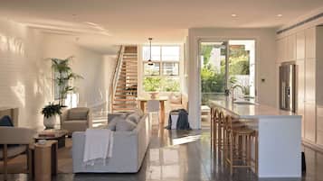 A modern kitchen with a central island, a dining area with a sofa and chairs, a staircase leading to an upper level, and a large sliding glass door to a garden.
