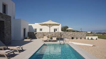 A modern pool area with lounge chairs and umbrellas, a stone wall, and a view of the landscape.