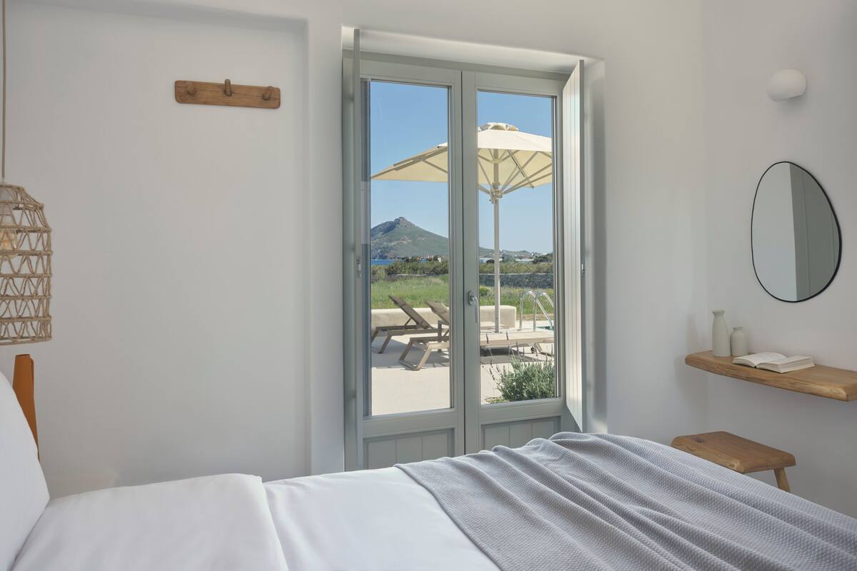 Un dormitorio con una cama, un ventanal con vista a una piscina y montañas, un estante de madera con libros, y un espejo montado en la pared.