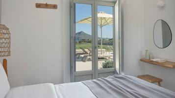 A bedroom with a bed, a window with a view of a pool and mountains, a wooden shelf with books, and a wall-mounted mirror.