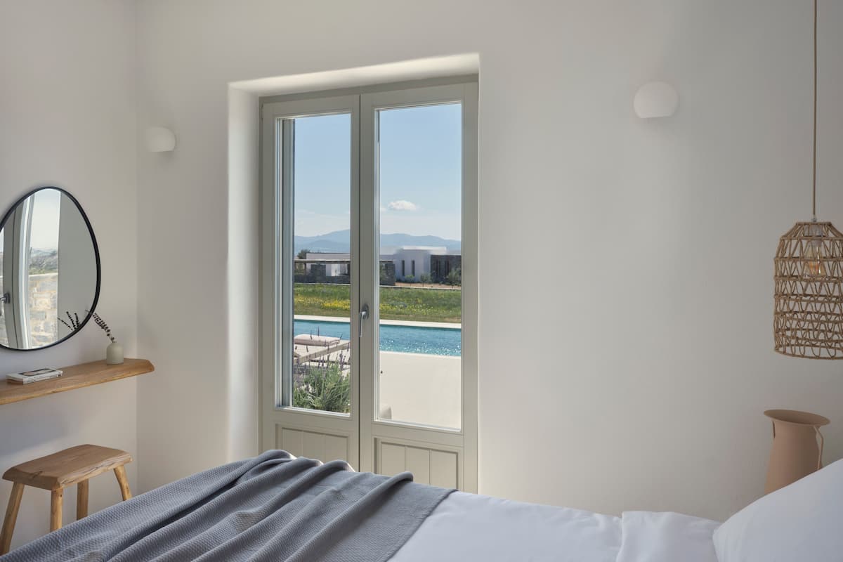 Un dormitorio con una cama, un taburete de madera, un espejo redondo y vistas a una piscina y montañas a través de un amplio ventanal.