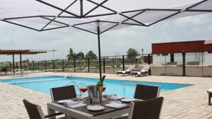 A poolside area with a table set for dining, a covered seating area, and a view of a building in the background. - Pride Elite Phaltan (Phaltan)