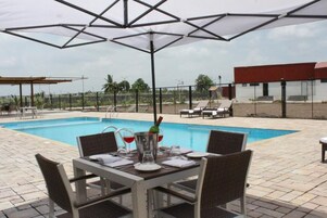 Zona de piscina con mesa dispuesta para comer, área de estar cubierta y vista a un edificio al fondo.