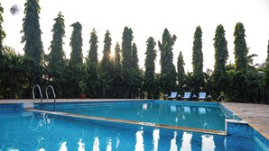 A swimming pool with clear blue water, surrounded by tall trees and a tiled deck with lounge chairs. - Pride Elite Phaltan (Phaltan)