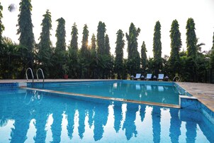 Una piscina con agua azul cristalina, rodeada de árboles altos y un deck de cerámica con sillas de descanso.