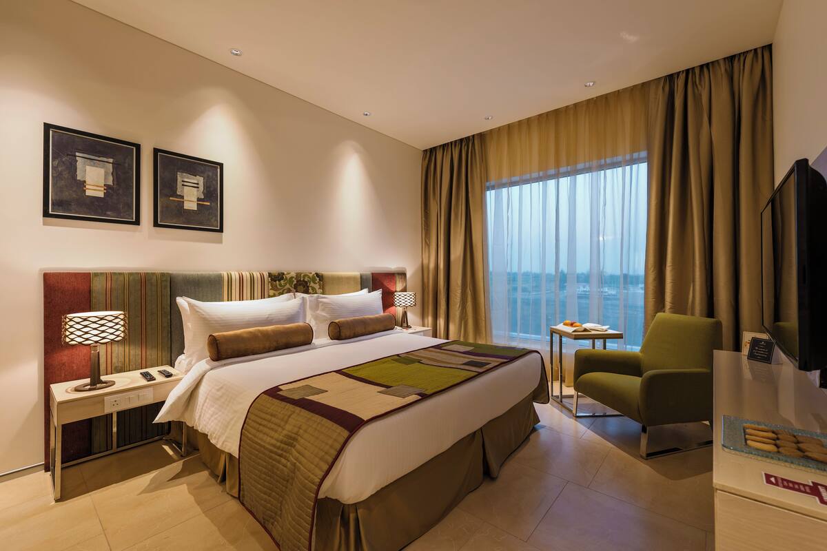Quarto de hotel com uma cama grande, uma escrivaninha com luminária, uma cadeira, uma televisão e uma janela com cortinas.