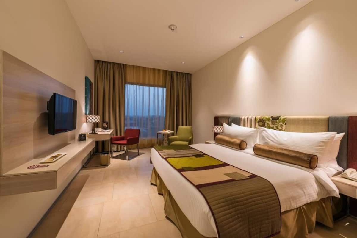 Quarto de hotel moderno com uma cama grande, uma escrivaninha, uma cadeira, uma TV e uma janela com cortinas.
