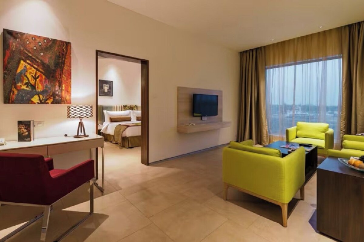 A modern hotel room with a desk, red chair, yellow sofa, and a TV on the wall.