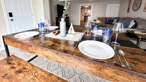 Un comedor con una mesa de madera preparada para cuatro personas, un florero con planta y vista del salón en el fondo.