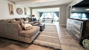 Una sala de estar con un sofá seccional, una mesa de centro, un televisor y un comedor al fondo.