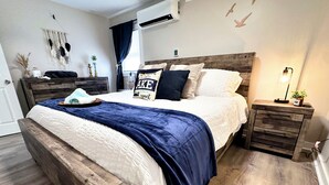 Un dormitorio con estructura de cama de madera, ropa de cama blanca, una manta de color azul oscuro y una mesita de noche con lámpara.