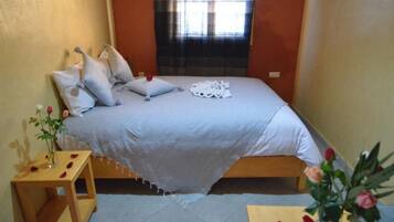 Una cama bien hecha con almohadas y una manta, una mesita de madera con un florero y una ventana con cortinas.