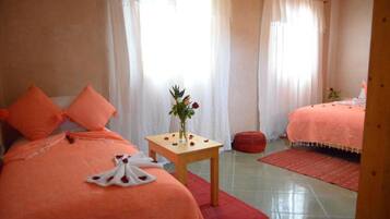 A bedroom with two beds, a small table with a vase of flowers, and a red ottoman.