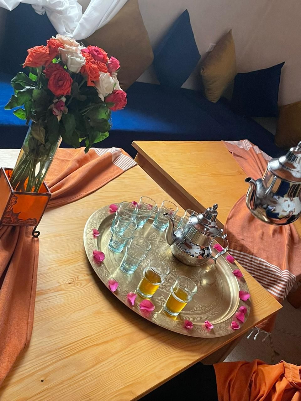 Una mesa de madera con una bandeja que sostiene una tetera y vasos, un florero con flores, y un cojín en un sofá azul al fondo.