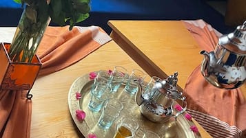 Una mesa de madera con una bandeja que sostiene una tetera y vasos, un florero con flores, y un cojín en un sofá azul al fondo.