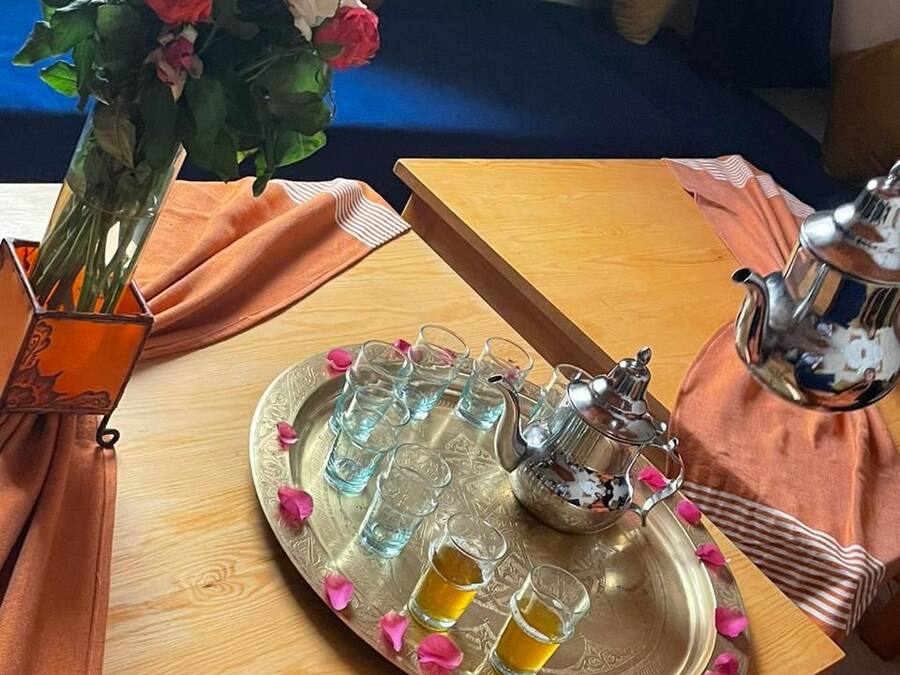 Una mesa de madera con una bandeja que sostiene una tetera y vasos, un florero con flores, y un cojín en un sofá azul al fondo.