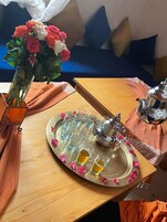 Una mesa de madera con una bandeja que sostiene una tetera y vasos, un florero con flores, y un cojín en un sofá azul al fondo.